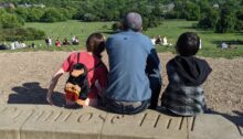 A man and his 2 daughters sit atop Primrose Hill, viewing the London skyline.