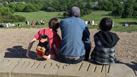 A man and his 2 daughters sit atop Primrose Hill, viewing the London skyline.